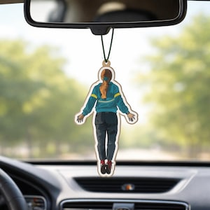May include: A car air freshener featuring a cartoon illustration of a person in a blue jacket and dark pants. The air freshener is hanging from a car's rearview mirror, with a green string. The background is a blurred view of trees.