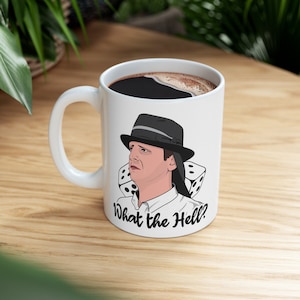 May include: White ceramic coffee mug with a cartoon illustration of a person wearing a hat and a surprised expression. The person is surrounded by dice. The text "What the Hell?" is printed on the mug.