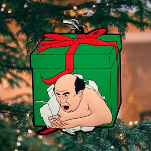 May include: A green gift box with a red bow and a man's face peeking out of the top. The man is wearing a white shirt and has a surprised expression on his face.
