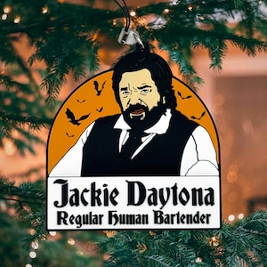 May include: A black and orange Halloween-themed ornament featuring a cartoon illustration of a man with a beard and the text "Jackie Daytona Regular Human Bartender".