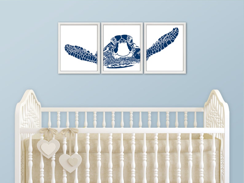 May include: Three white framed prints of a blue silhouette of a sea turtle. The prints are arranged in a row on a light blue wall above a white crib with a beige mattress and white heart-shaped decorations.