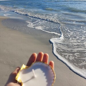May include: A white candle in a gold-trimmed seashell, held in front of a beach scene. The candle has a single wick. The seashell is white with a gold edge. The background shows the ocean and a sandy beach.