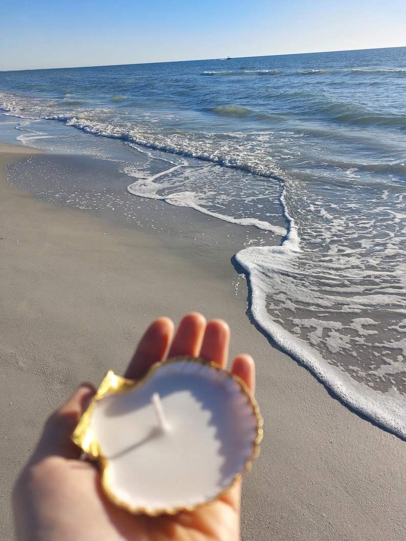 May include: A white candle in a gold-trimmed seashell, held in front of a beach scene. The candle has a single wick. The seashell is white with a gold edge. The background shows the ocean and a sandy beach.
