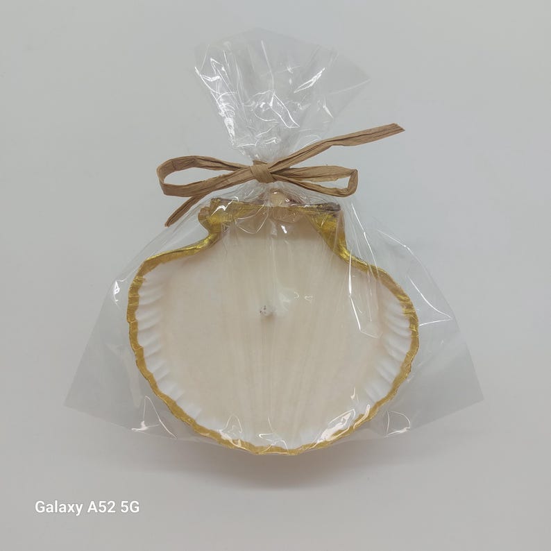 May include: A seashell-shaped candle with a gold-painted rim, packaged in clear cellophane and tied with a raffia bow. The candle is off-white and has a single wick. The overall aesthetic is coastal and decorative.