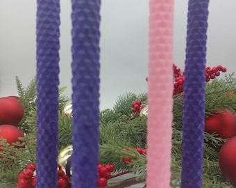 May include: Four Advent candles in a gold metal holder. Two purple candles and one pink candle are visible. The candles are decorated with a honeycomb pattern. The candles are surrounded by green pine branches and red berries.