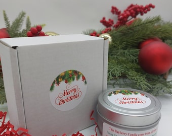 May include: A silver tin candle with a red and green Christmas-themed label that reads "Merry Christmas". The candle is sitting on a red shredded paper with a white box behind it. The box also has a red and green Christmas-themed label that reads "Merry Christmas".