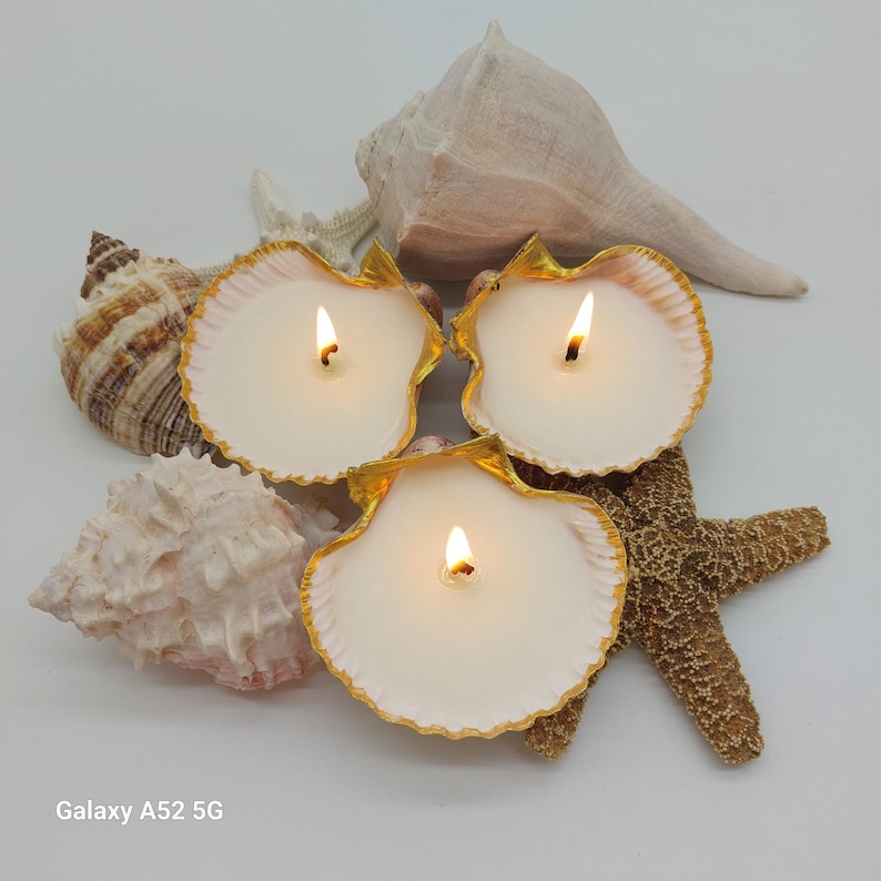 May include: Three lit seashell candles with gold trim, surrounded by seashells and a starfish. The candles have a white wax and are set against a white background, creating a coastal theme.