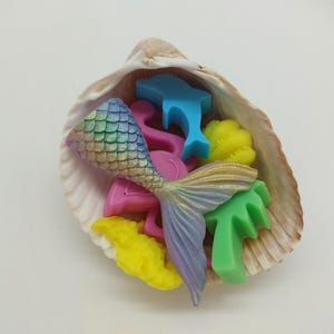 May include: A seashell filled with colorful, small, decorative items. The items include a mermaid tail, dolphins, flamingos, palm trees, and coral. The mermaid tail has a scale pattern and is iridescent.