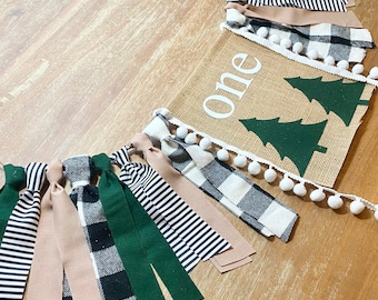 May include: A burlap banner with the word "one" in white letters and two green felt Christmas trees. The banner is decorated with white pom-poms and a string of fabric scraps in black and white buffalo plaid, green, and tan.