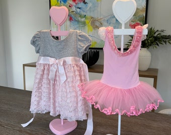 May include: Two pink dresses on heart-shaped display stands. The dress on the left is a grey and pink layered dress with a white ribbon. The dress on the right is a pink tulle tutu with pink ribbon trim.