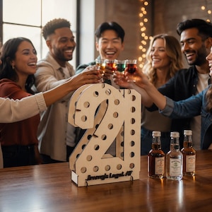 May include: A light-colored wooden number 21 shot glass holder with a series of circular cutouts and the text Jimmy's Legal AF printed on the base, accompanied by three small glass bottles of liquor placed on a wooden surface.