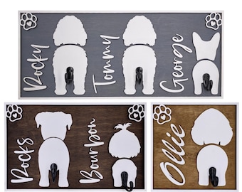 May include: Three wooden dog leash hooks with white silhouettes of dogs' rear ends and their names, "Rocky", "Tommy", and "George", written vertically. The hooks are painted gray with white paw prints and hearts.