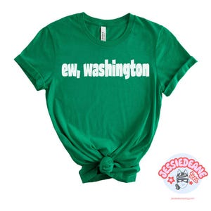 May include: A bright green short-sleeved t-shirt featuring the white text "ew, washington" printed across the chest in a bold, sans-serif font. The garment has a crew neckline and is shown with the bottom hem tied into a small knot at the center.