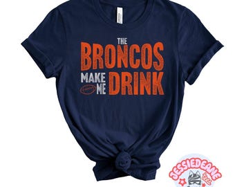 May include: A navy blue short-sleeved t-shirt featuring the orange and white text THE BRONCOS MAKE ME DRINK with a small graphic of an American football. The shirt has a crew neckline and a knotted hem detail at the bottom centre of the garment.