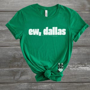 May include: A green t-shirt with the white text "ew, dallas". The shirt has a round neck and short sleeves. A small graphic of a masked character is on the lower right side of the shirt.