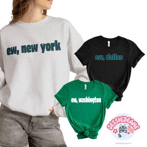 May include: A light gray crewneck sweatshirt with the dark teal text "ew, new york", a black t-shirt with the teal text "ew, dallas", and a green t-shirt with the white text "ew, washington" printed across the chest of each garment.