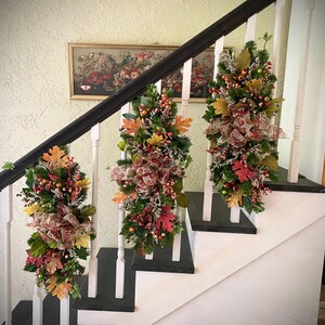 Puede incluir: Tres guirnaldas decorativas con temática otoñal adornan una escalera. Cada guirnalda presenta una mezcla de follaje verde, bayas rojas y coloridas hojas de otoño. Cintas estampadas marrones y rojas añaden un toque festivo. La escalera tiene balaustres blancos y peldaños negros.