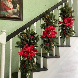 May include: Three Christmas garlands with red bows and gold accents hang on a white staircase railing. The garlands are made of green pine branches, red berries, and gold pine cones.