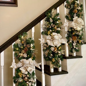 May include: A white staircase decorated with three gold and white Christmas garlands with lights. Each garland has a large gold and white bow.