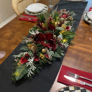 May include: A black table runner with a Christmas centerpiece featuring red and gold ornaments, pine branches, and red flowers. The centerpiece is on a wooden table with white and black checkered plates and red napkins.