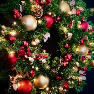 May include: A Christmas wreath decorated with red and gold ornaments, pine branches, and red berries. The wreath is lit with white fairy lights.