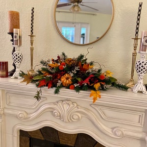 Puede incluir: Una chimenea blanca con un diseño decorativo tallado. Un centro de mesa de otoño con calabazas, bayas y hojas se encuentra en la repisa de la chimenea. Dos candelabros dorados con velas a cuadros negros y blancos están a cada lado del centro de mesa.