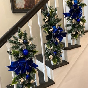 Op de afbeelding: Drie blauwe en zilveren kerstkransen met blauwe strikken en witte bessen hangen aan een witte leuning.