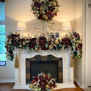 May include: A decorated fireplace mantel with a large burgundy and gold wreath, garland, and floral arrangement. Two lamps flank the mantel, and two deer figurines and a framed photo are on top. The fireplace has a white surround and a stone facade.