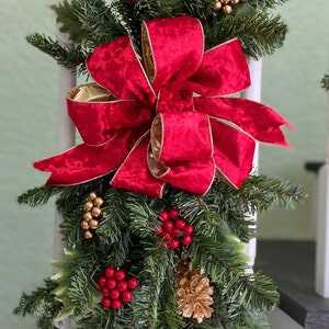 May include: A red velvet bow with gold trim decorates a green garland with gold pine cones and red berries. The garland is hanging on a white railing.