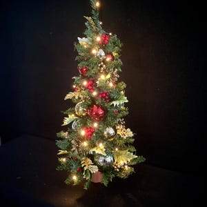 May include: A miniature artificial Christmas tree with green pine needles, illuminated by small warm white string lights. The tree is decorated with shiny red and silver baubles, clusters of red berries, and gold-toned decorative leaves.