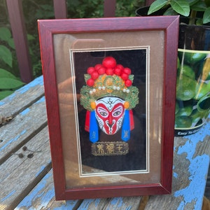 May include: A framed artwork featuring a colorful Chinese opera mask with red, green, blue, and gold accents. The mask is set against a black background and has the text "青猴王" written in gold below it.
