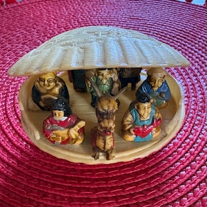 May include: A circular cream-colored figurine featuring a textured roof structure sheltering seven small painted figures and a central dragon. The figures are depicted in various poses, wearing colorful robes, and are arranged on a flat base.