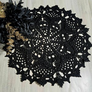 May include: A black, crocheted doily with a scalloped edge. The intricate openwork design features a central floral pattern and a decorative border. The doily is displayed on a light-colored wooden surface, with black and gold decorative elements.