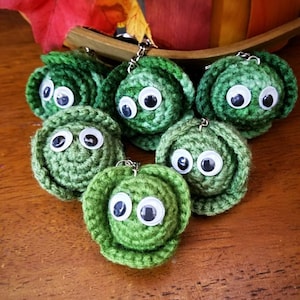 May include: A collection of six handmade green crochet cabbage keychains with googly eyes. Each keychain features a silver-colored metal ring for attaching to keys or bags. The keychains are arranged on a wooden surface.