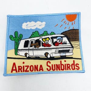 May include: A blue and yellow embroidered patch with a white camper van driving on a yellow road. The van has three people inside and a cactus and a sun in the background. The text "Arizona Sunbirds" is at the bottom of the patch.