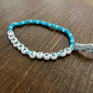 May include: A blue and white beaded bracelet with the words "COWBOY LIKE ME" spelled out on the beads. A silver charm in the shape of a boot with a floral design is attached to the bracelet.