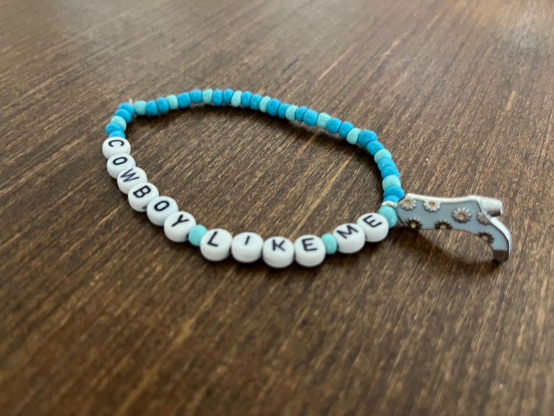 May include: A blue and white beaded bracelet with the words "COWBOY LIKE ME" spelled out on the beads. A silver charm in the shape of a boot with a floral design is attached to the bracelet.