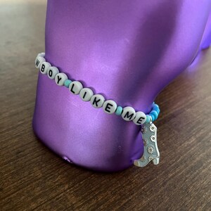 May include: A white beaded bracelet with the words "BOY LIKE ME" spelled out in black letters. The bracelet has blue and white beads and a silver charm in the shape of a boot.
