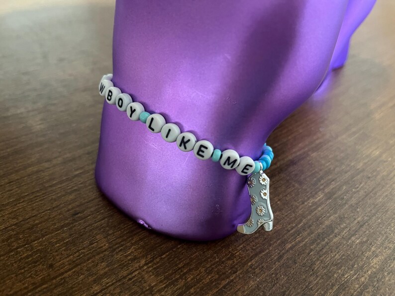 May include: A white beaded bracelet with the words "BOY LIKE ME" spelled out in black letters. The bracelet has blue and white beads and a silver charm in the shape of a boot.
