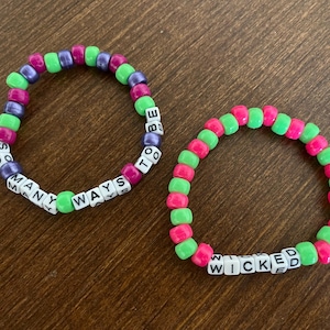 Puede incluir: Dos pulseras de cuentas. Una pulsera tiene cuentas verdes, moradas y rosas con las palabras "MANY WAYS TO BE" escritas. La otra pulsera tiene cuentas verdes y rosas con la palabra "WICKED" escrita.