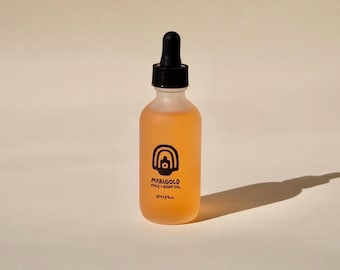 May include: A frosted glass bottle with a black dropper top, filled with orange liquid. The bottle has a black and white label that reads "Marigold Face + Body Oil" and "2 fl oz / 59 ml". The bottle casts a shadow on a neutral background.