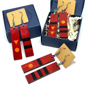 May include: Red rectangular wooden earrings with black stripes and orange and yellow geometric shapes. The earrings are displayed in blue gift boxes with kraft paper tags. The earrings are on a white background.
