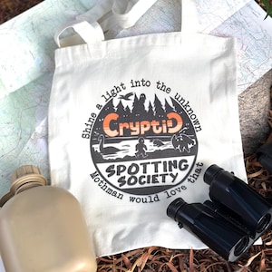 May include: Cream-colored tote bag with the words "Cryptid Spotting Society" in a circular design. The design includes illustrations of cryptids and the text "Shine a light into the unknown". A canteen and binoculars are also visible.