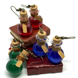 May include: Earrings featuring miniature glass potion bottles in red, blue, and green, each with a cork stopper and a small tag. The earrings are displayed on a small, dark red book.