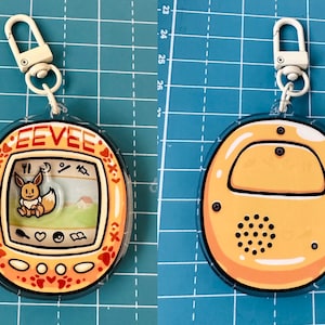 May include: A yellow and orange plastic keychain with a clear plastic cover. The keychain features a digital pet toy with a cartoon image of Eevee, a Pokemon character, on the screen. The text "EEVEE" is printed on the toy. The back of the keychain is plain yellow.
