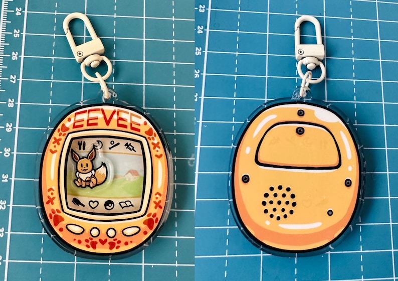 May include: A yellow and orange plastic keychain with a clear plastic cover. The keychain features a digital pet toy with a cartoon image of Eevee, a Pokemon character, on the screen. The text "EEVEE" is printed on the toy. The back of the keychain is plain yellow.