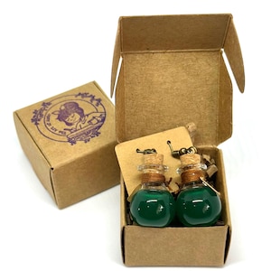 May include: Two small glass potion bottles filled with green liquid, each with a cork stopper and a metal hook, presented in a brown cardboard box. A second box with a purple stamp is visible.