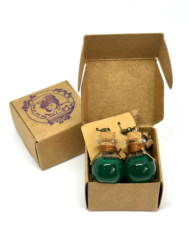 May include: Two small glass potion bottles filled with green liquid, each with a cork stopper and a metal hook, presented in a brown cardboard box. A second box with a purple stamp is visible.