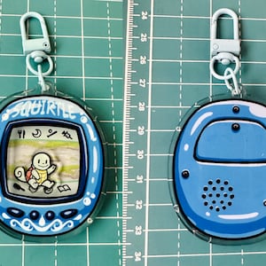 May include: A blue and white plastic keychain featuring a Squirtle character from the Pokemon franchise. The keychain is shaped like a Tamagotchi and has a clear plastic case. The Squirtle character is visible on the screen, and the keychain has a blue and white design.