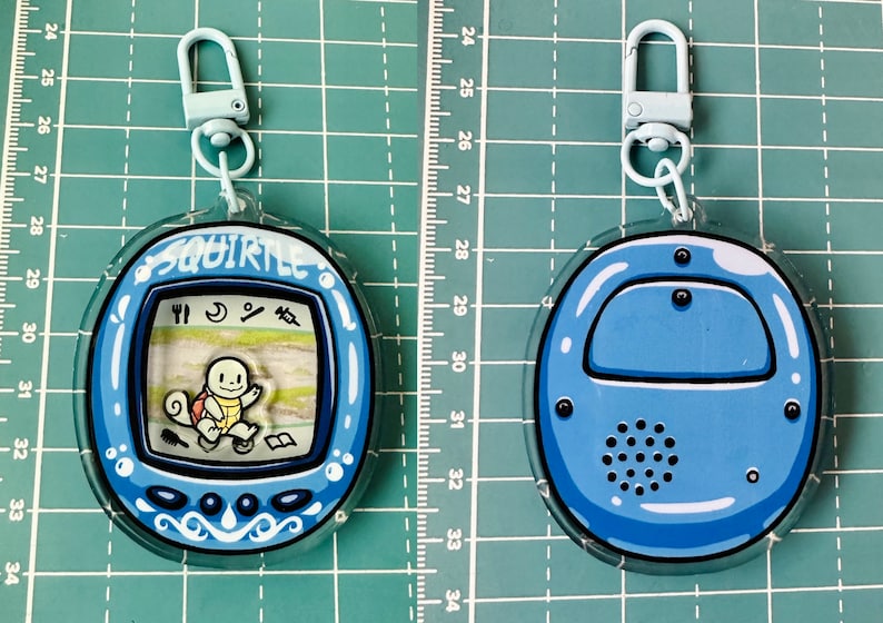 May include: A blue and white plastic keychain featuring a Squirtle character from the Pokemon franchise. The keychain is shaped like a Tamagotchi and has a clear plastic case. The Squirtle character is visible on the screen, and the keychain has a blue and white design.